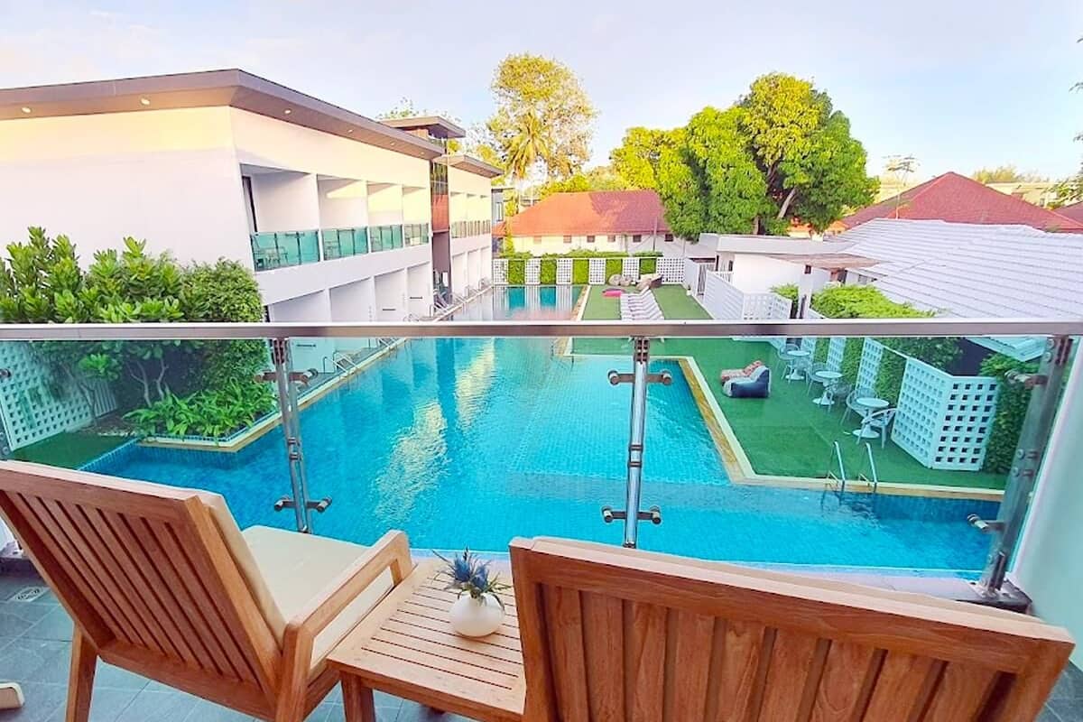 A poolside area with two wooden chairs and a small table, overlooking a swimming pool and a garden area with white fences and trees.