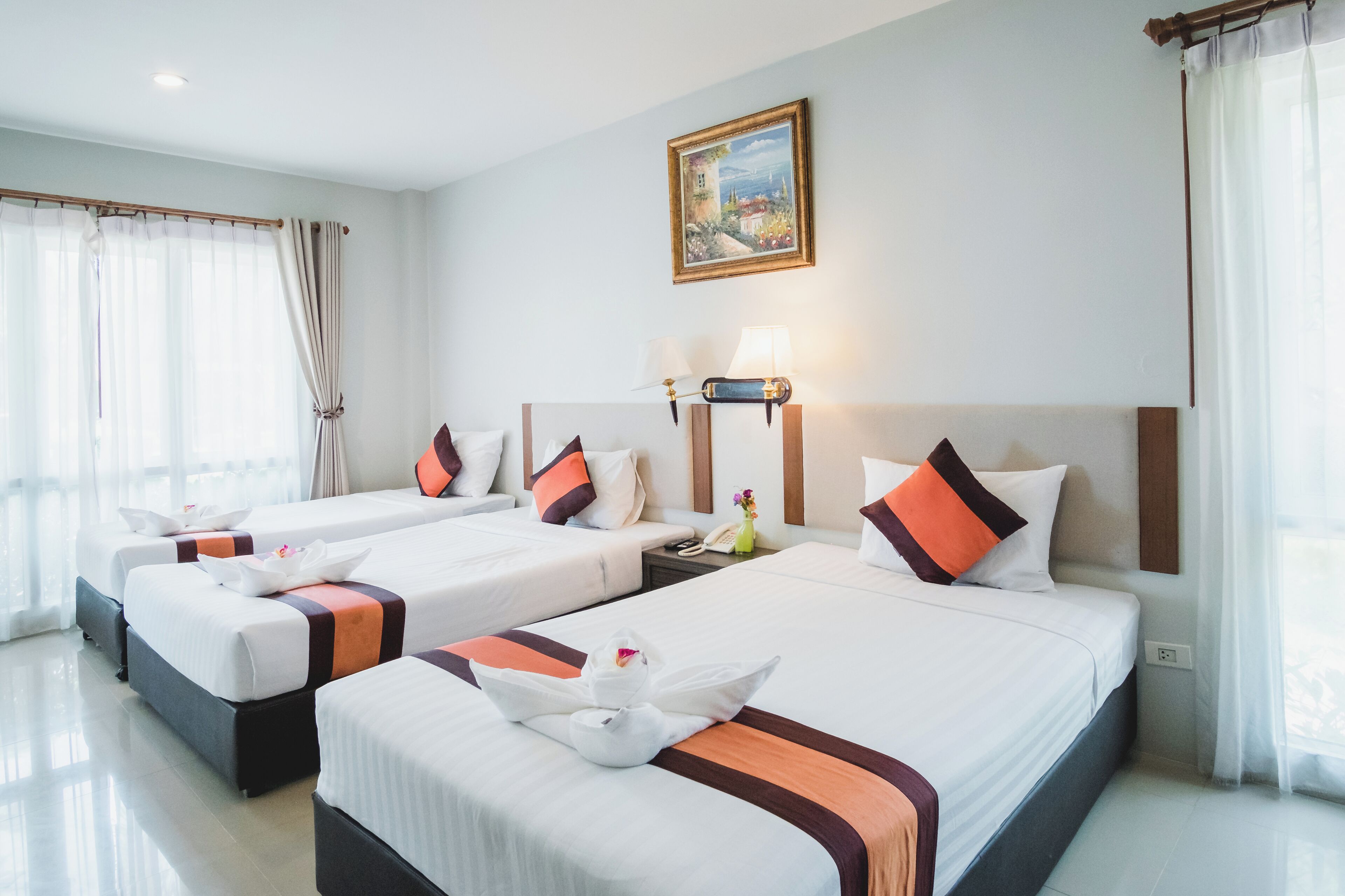 Habitación de hotel con tres camas, cada una con ropa de cama blanca y detalles en naranja, un cuadro en la pared y una lámpara en la mesita de noche.