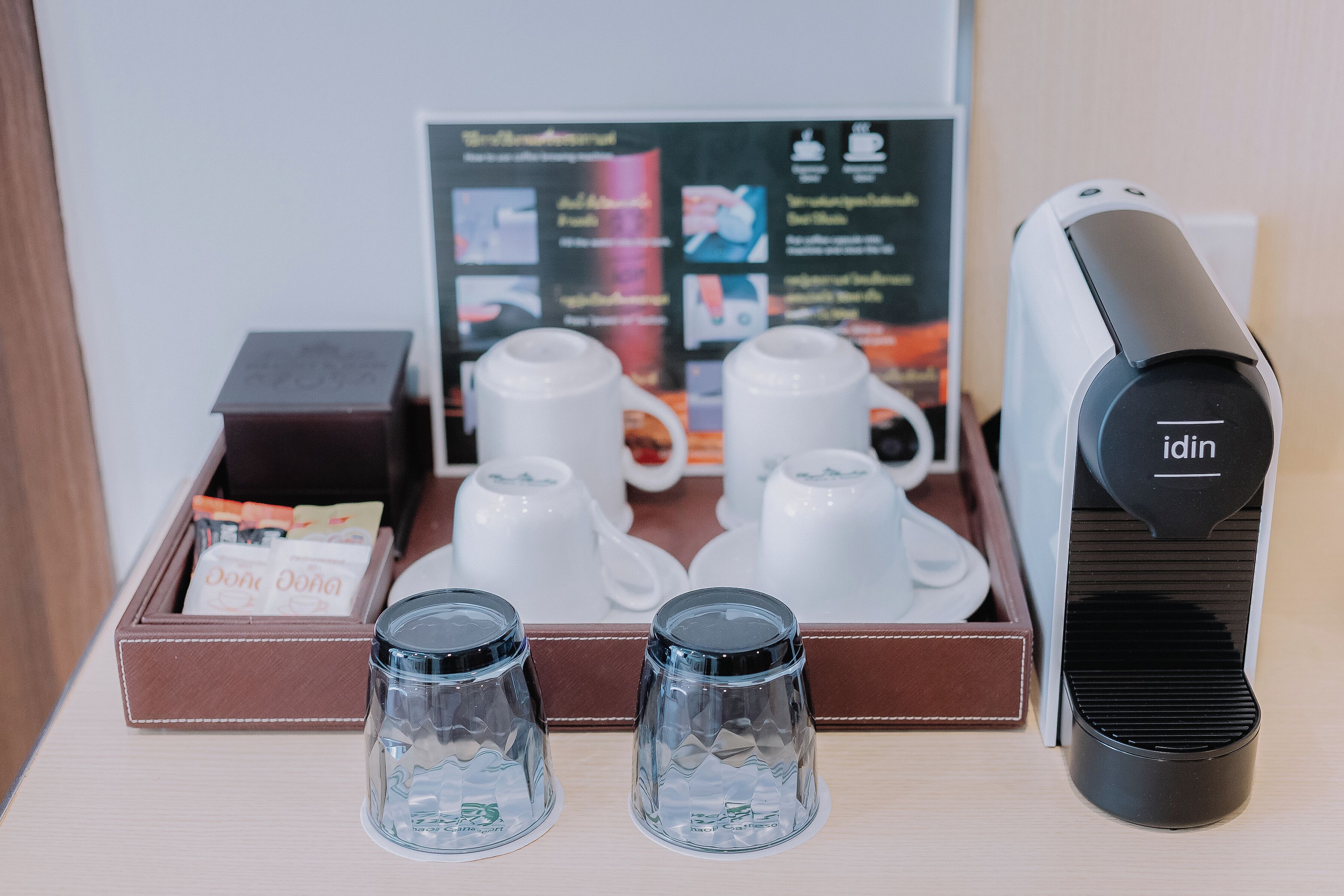Une machine à café, deux tasses en verre, un plateau avec une boîte de sachets de thé et un tableau affichant le menu dans une chambre d’hôtel.