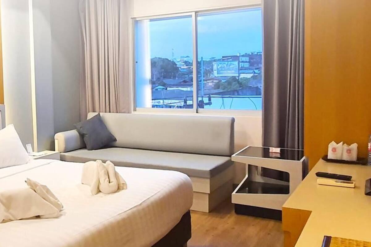 A hotel room with a large bed, a sofa, a desk with a computer, and a window with a city view.