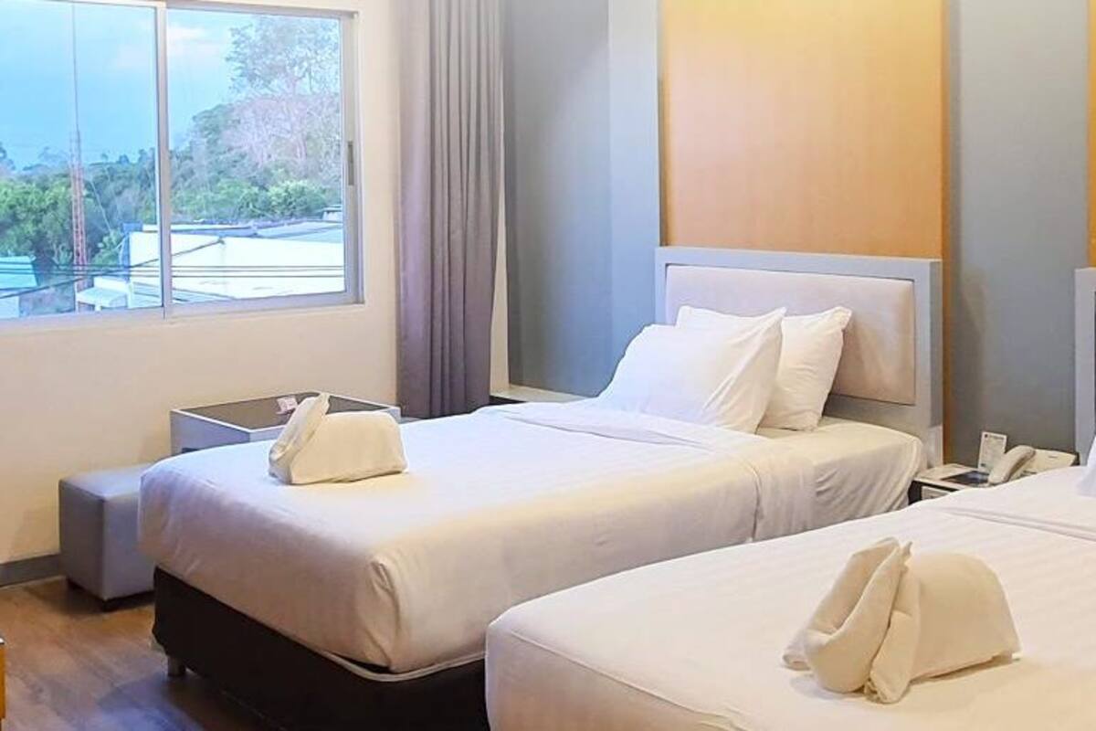 A hotel room with two beds, a desk, a chair, and a window with a view of greenery.