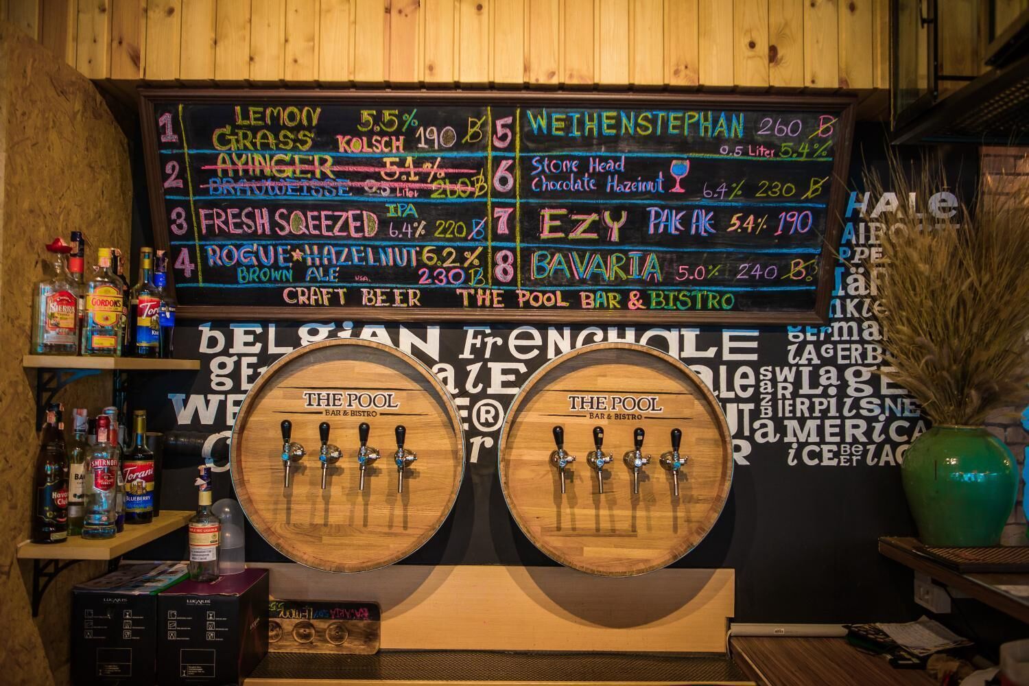 Un bar avec un tableau noir servant de menu, où sont indiquées les différentes bières proposées et leurs prix. Devant le menu, on trouve deux tonneaux en bois équipés de robinets.