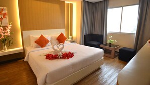 Ein Hotelzimmer mit einem großen Bett, weißer Bettwäsche, orangefarbenen Kissen, einer Handtuch-Herzanordnung, einem kleinen Tisch mit einer Pflanze und Blick ins Freie durch ein Fenster mit Vorhängen.
