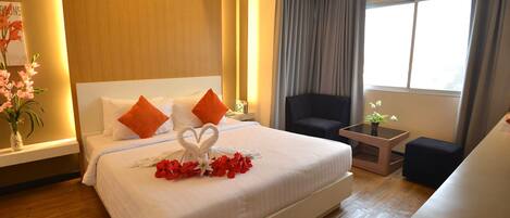 Habitación de hotel con una cama grande, ropa de cama blanca, almohadas naranjas, una disposición de toallas con forma de corazón, una mesita con una planta y vistas al exterior a través de una ventana con cortinas.
