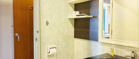 Un baño con puerta de madera, porta papel higiénico, espejo y lavamanos con encimera negra.
