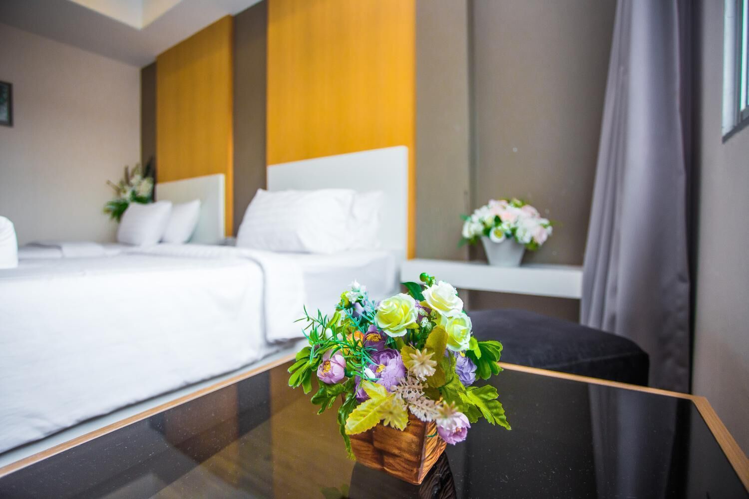 A hotel room with a bed, a bedside table with a floral arrangement, and a window with curtains.