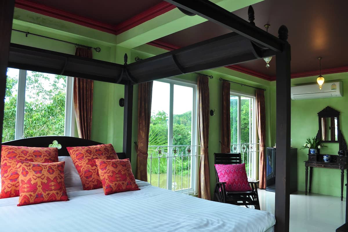 A four-poster bed with floral pillows, a view of greenery through large windows, and a balcony chair.