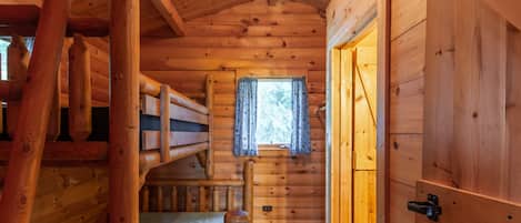 Interior de una cabaña de madera con una litera, una ventana con cortinas y una puerta.