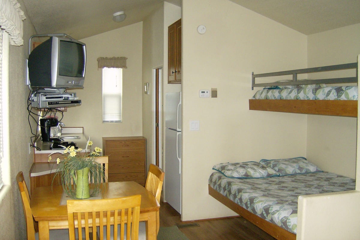 A compact room with a bunk bed, a small desk with a television, a refrigerator, and a dining area with a table and chairs.