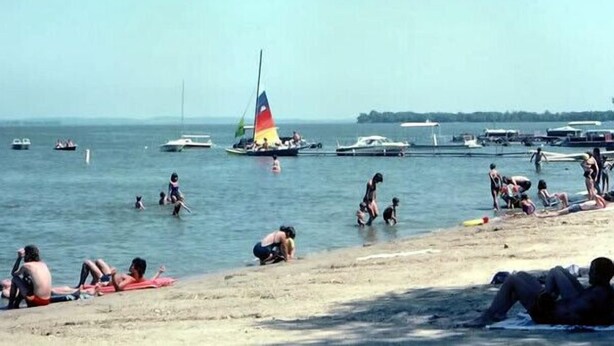 A beach with people sunbathing and swimming, boats in the water, and a sailboat with a colorful sail. - Lakeland RV Campground (Milton)