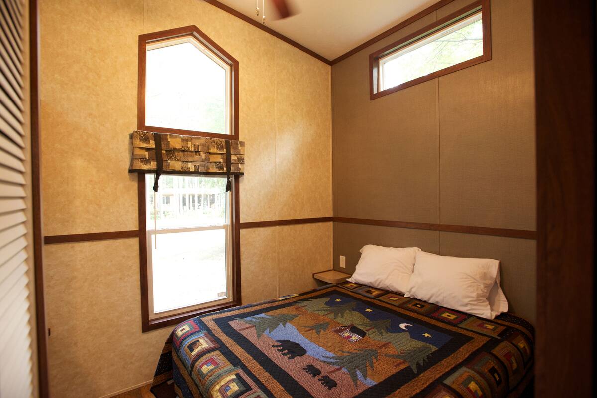 A bedroom with a large window, a quilt with a forest design, and a bed with white pillows.