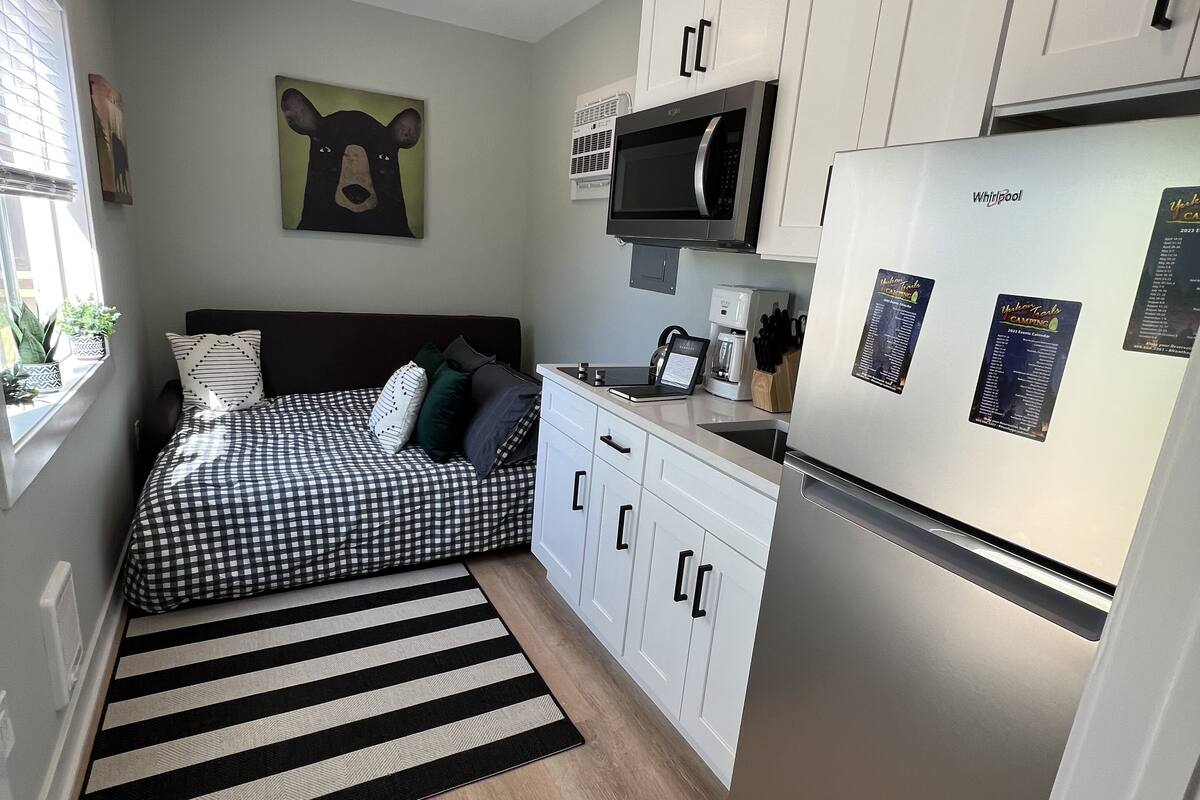A compact kitchen with a refrigerator, microwave, and coffee maker. There is a bed with a checkered bedspread and pillows. A black and white striped rug is on the floor.