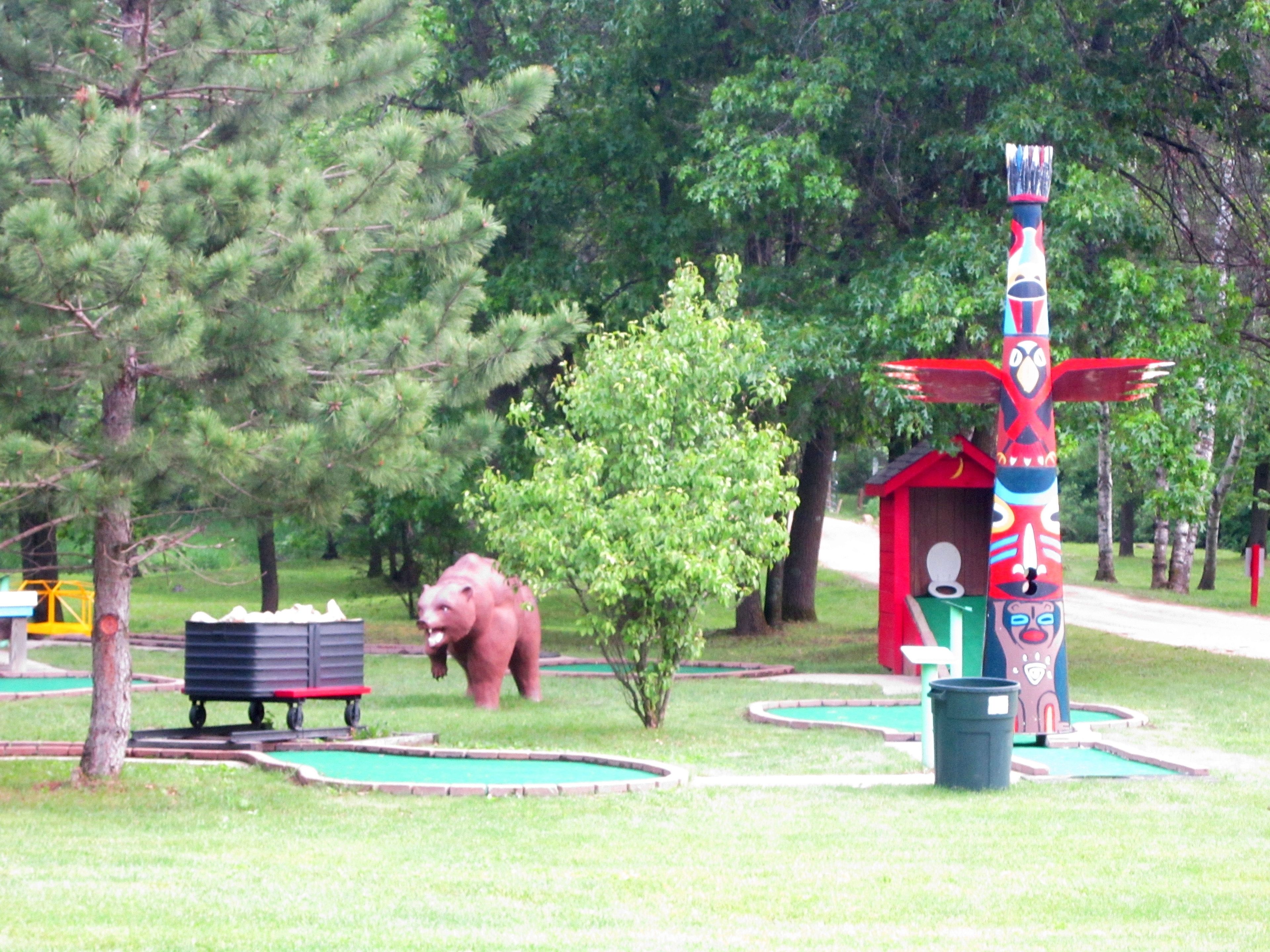 A miniature golf course with a bear statue, a totem pole, and a small red building.