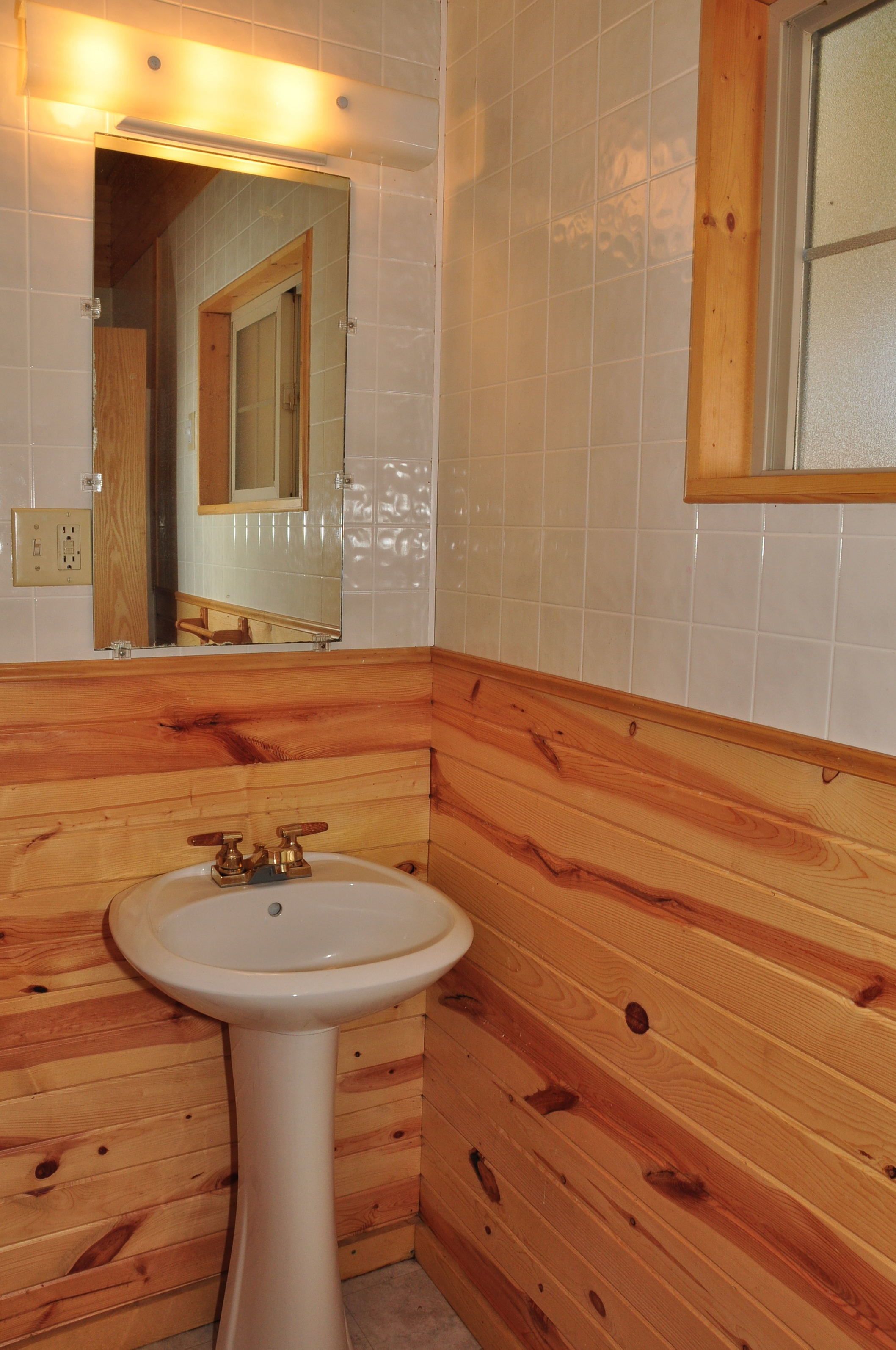 A bathroom with wooden paneling, a pedestal sink, and a mirror with a wooden frame.