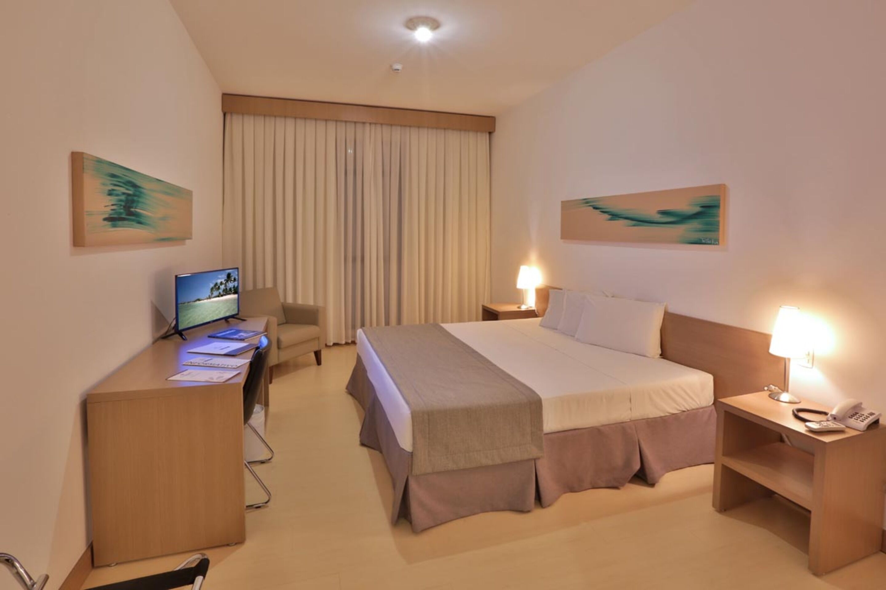 Une chambre d’hôtel avec un lit, des tables de chevet, un bureau avec un ordinateur, une chaise et des œuvres d’art accrochées aux murs.