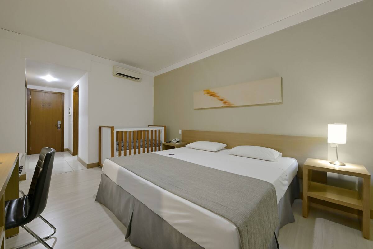 Ein Hotelzimmer mit einem großen Bett, einem Schreibtisch mit Stuhl, einem Nachttisch mit Lampe und einem Babybett.