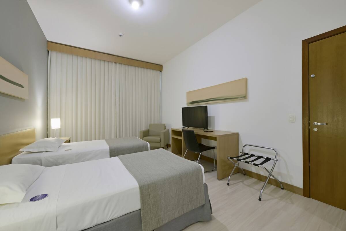 Ein Hotelzimmer mit zwei Betten, einem Schreibtisch mit Computer, einem Fernseher, einem Sessel und einer Tür.