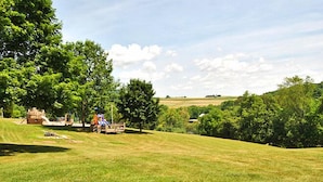 A grassy park area with playground equipment, trees, and a distant view of a field.