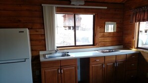 A compact kitchen with wooden cabinets, a white countertop, a sink, and a window with a view of a wooden building outside. - Sun Valley Camping and RV Resort (Narvon)