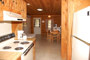 Interior de una cabaña de madera con una cocina que incluye estufa, refrigerador y una mesa de comedor con sillas.