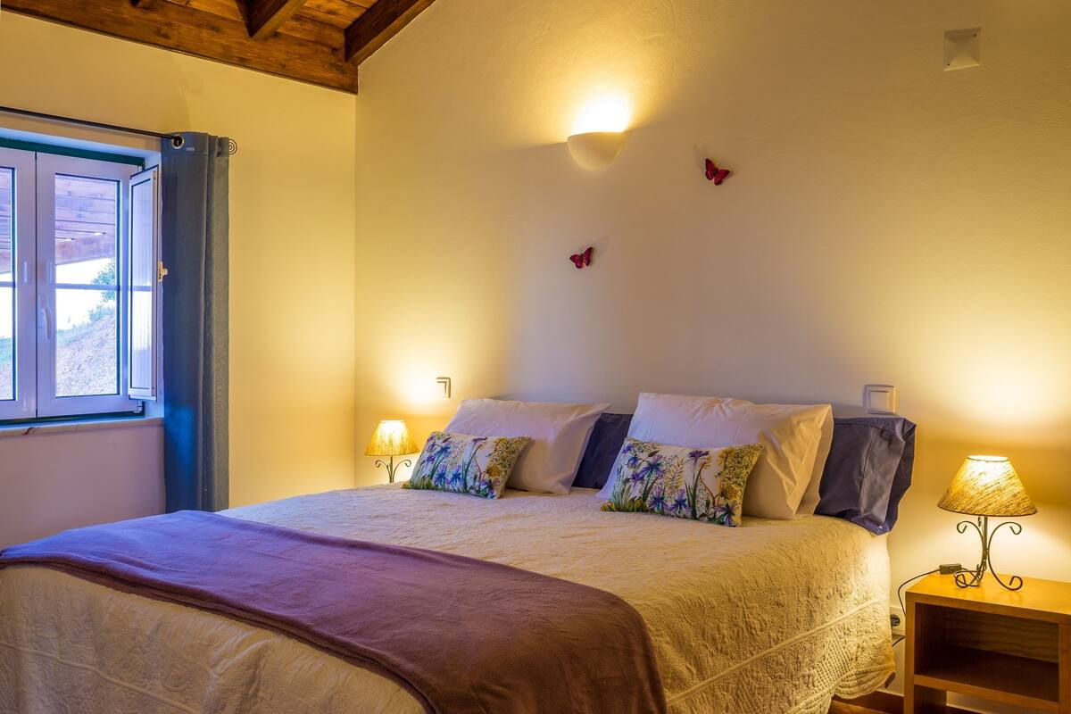 A bedroom with a bed, pillows, a nightstand with a lamp, and a window with curtains.