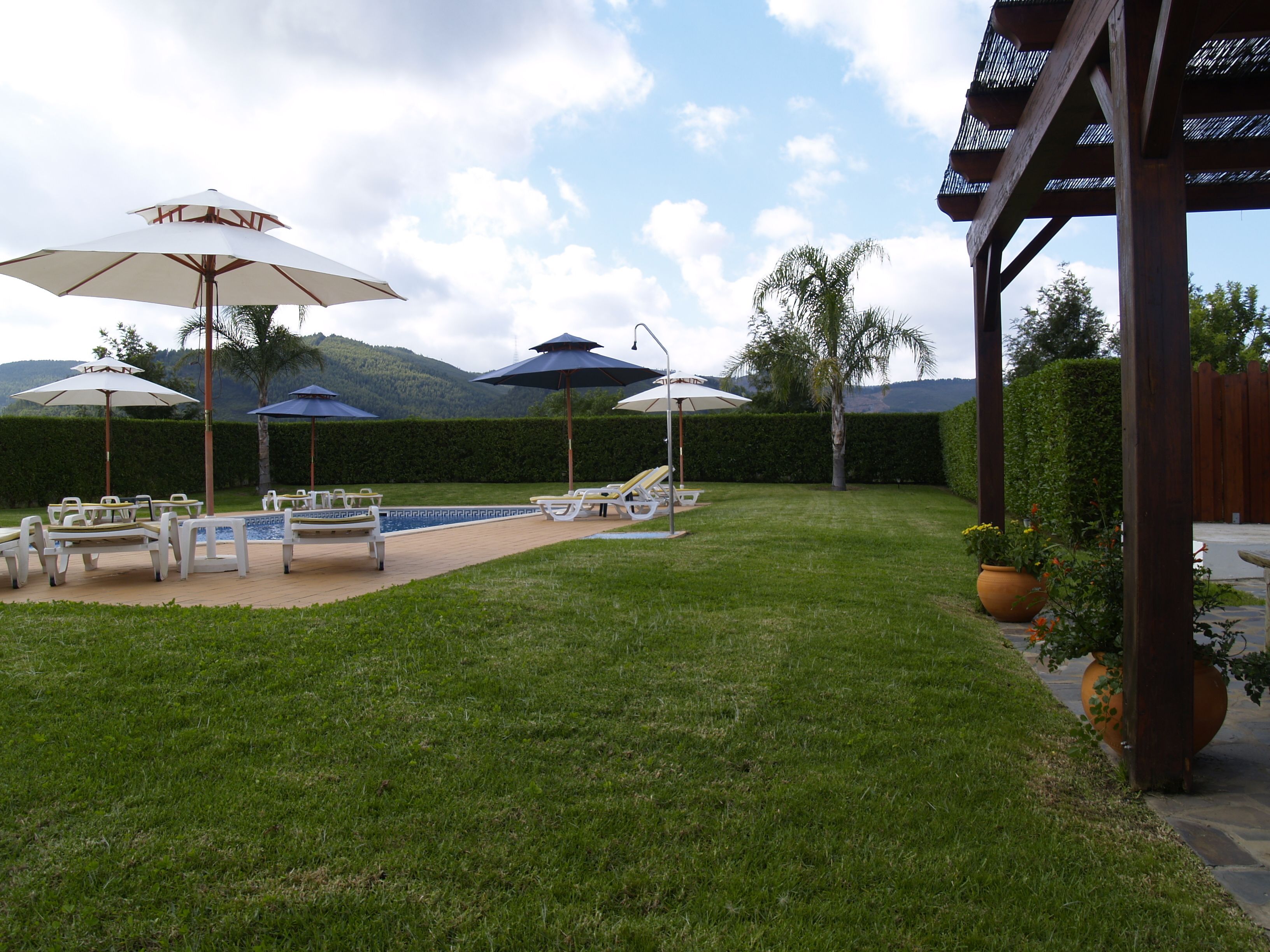 Un área junto a la piscina con sillones de descanso y sombrillas, rodeada por un césped cuidado y una pérgola de madera.