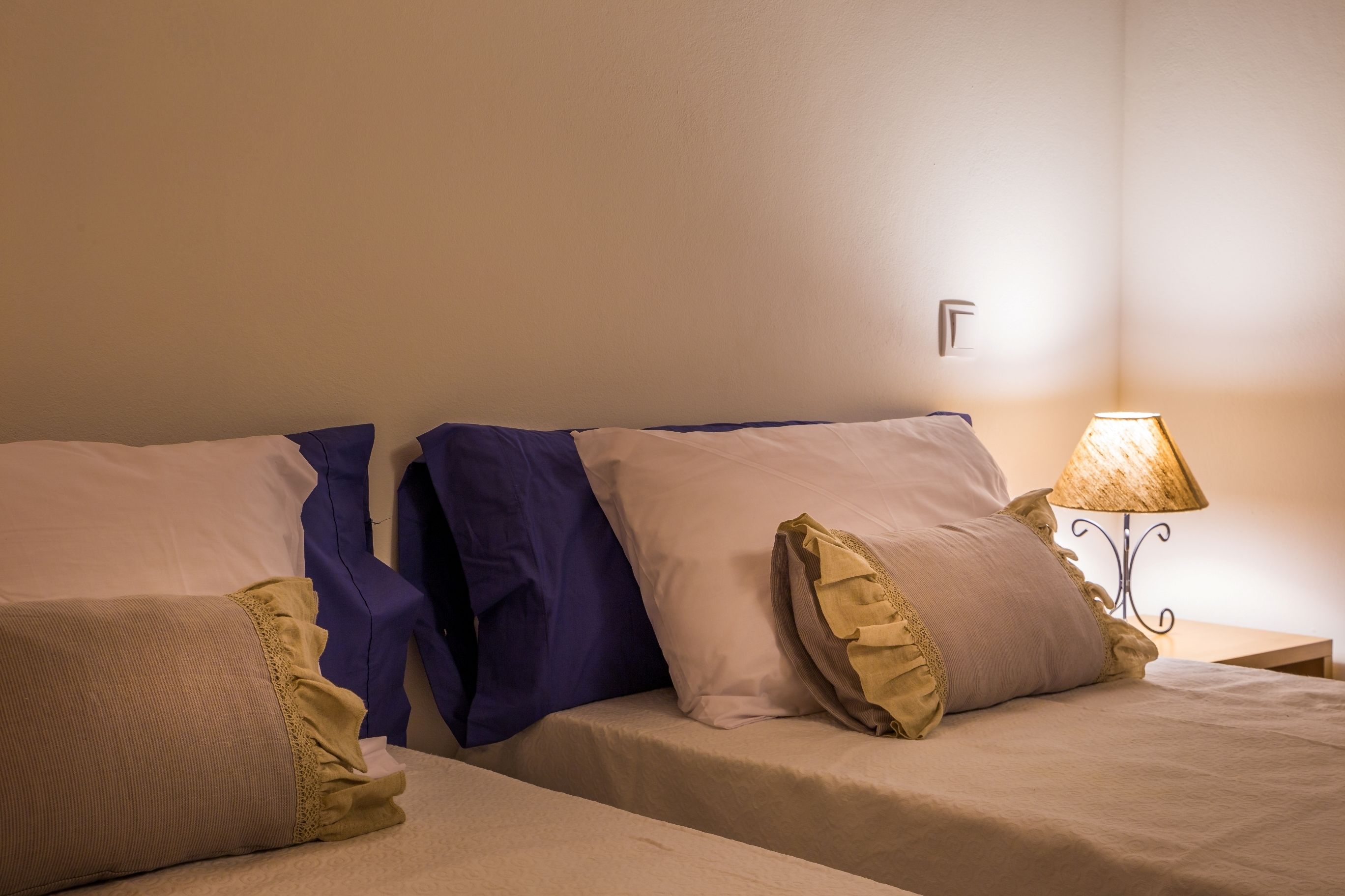 Ein Hotelzimmer mit zwei Betten, jedes mit einer Decke und Kissen in unterschiedlicher Farbe. Dazwischen steht ein Nachttisch mit einer Lampe.