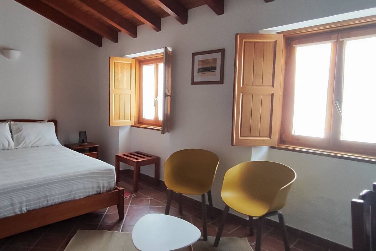 A bedroom with a bed, two chairs, a small table, a window with wooden shutters, and a picture on the wall.