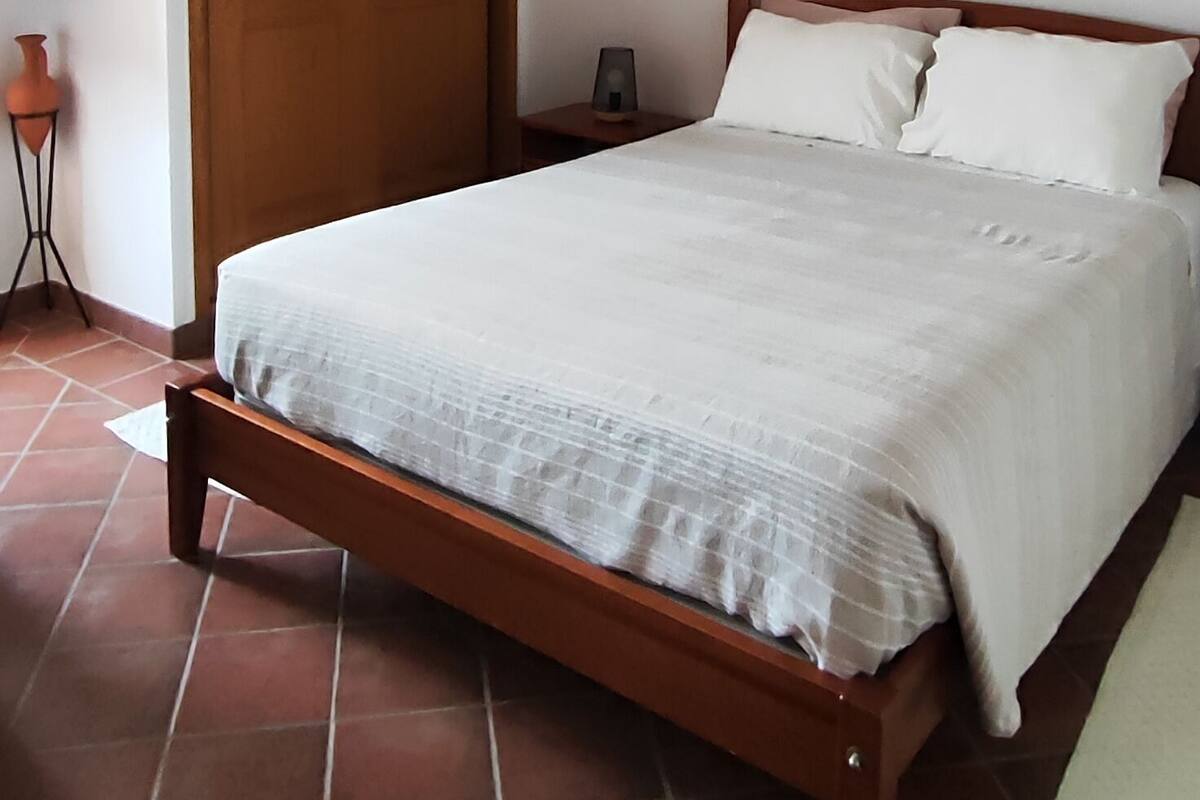 A bedroom with a wooden bed, a white wall, a wooden ceiling, and a tiled floor.
