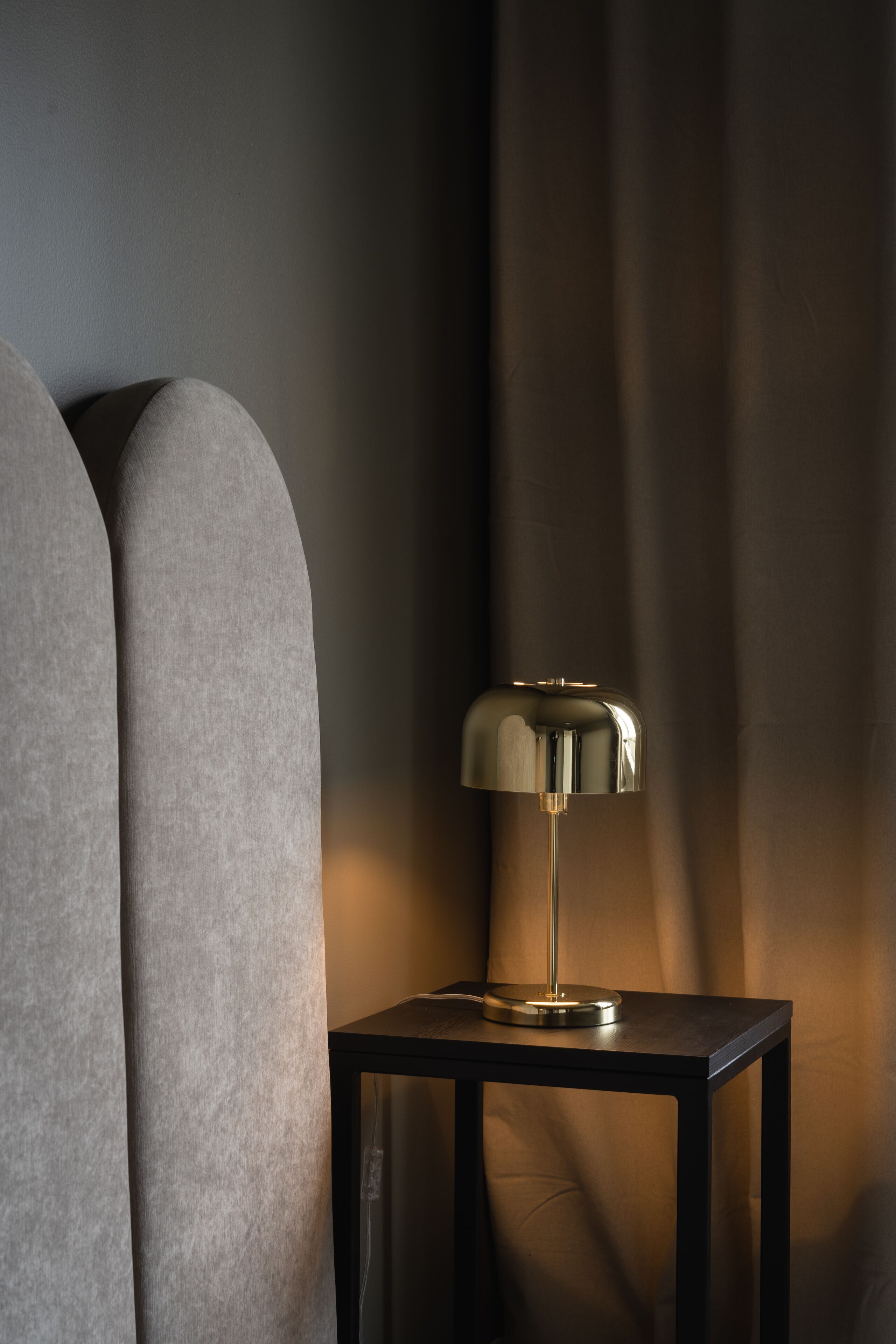Une lampe moderne posée sur une petite table à côté d’un canapé, avec un mur sombre à texture et un rideau en arrière-plan.