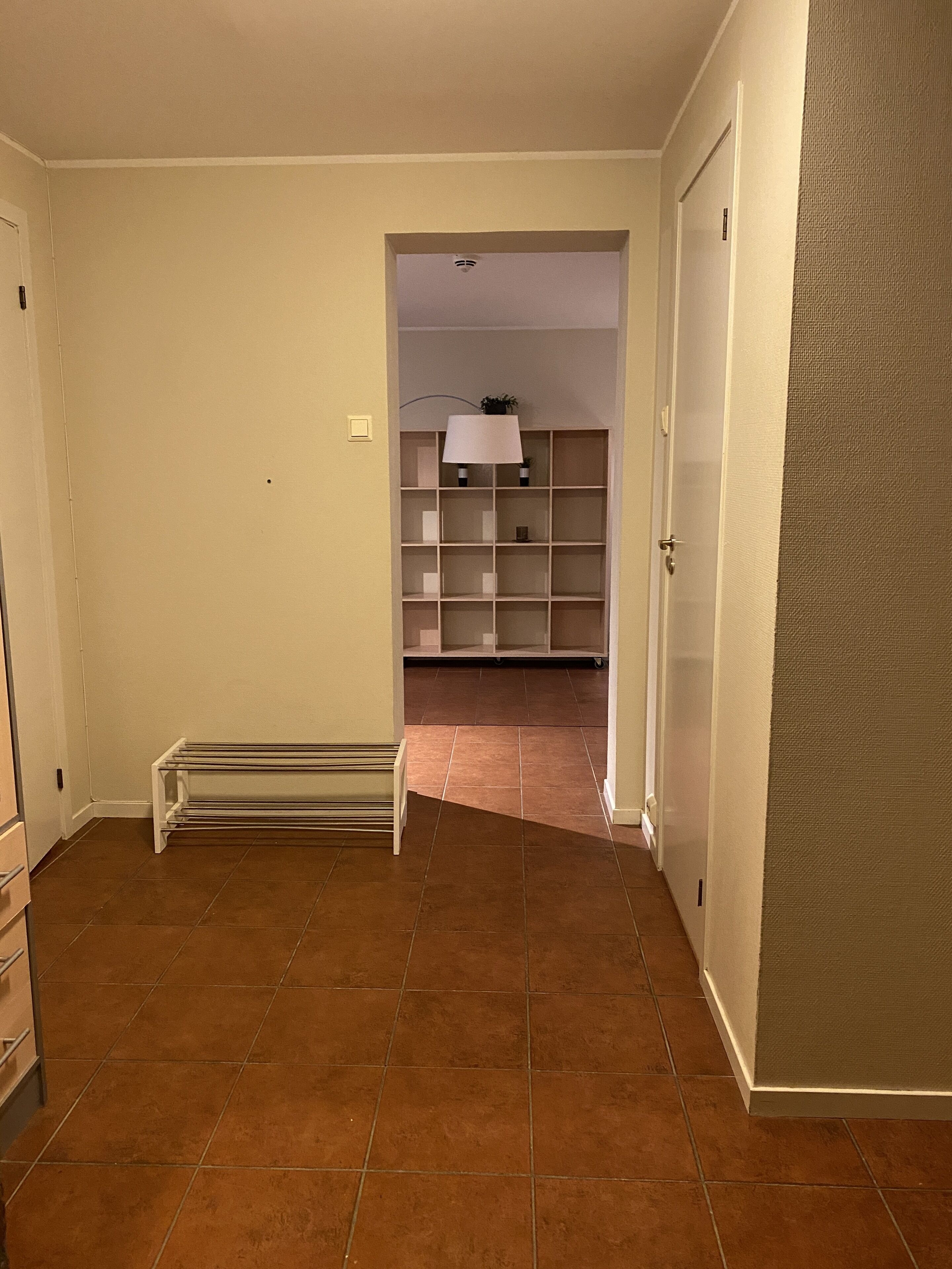 Un pasillo con suelo de baldosas, un radiador y una puerta que da a una habitación con estanterías y una lámpara.