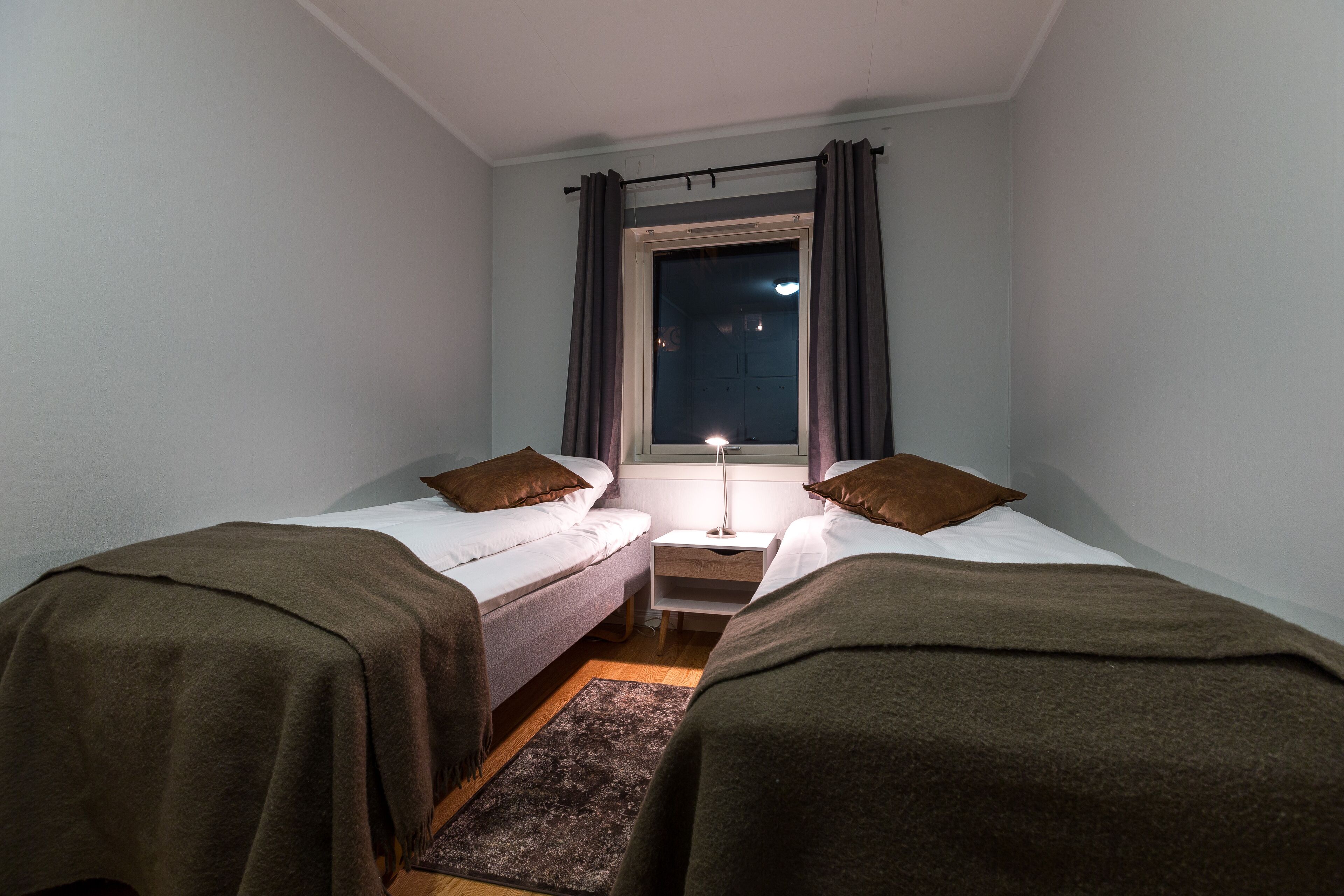 A small, dimly lit room with two single beds, a nightstand, and a window with curtains.