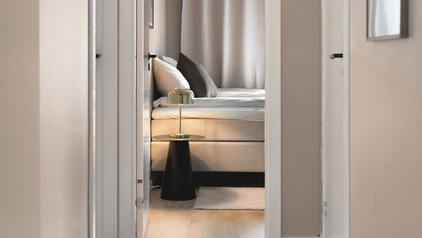 Un couloir étroit mène à une chambre à coucher qui contient un lit, une table de chevet et une commode.