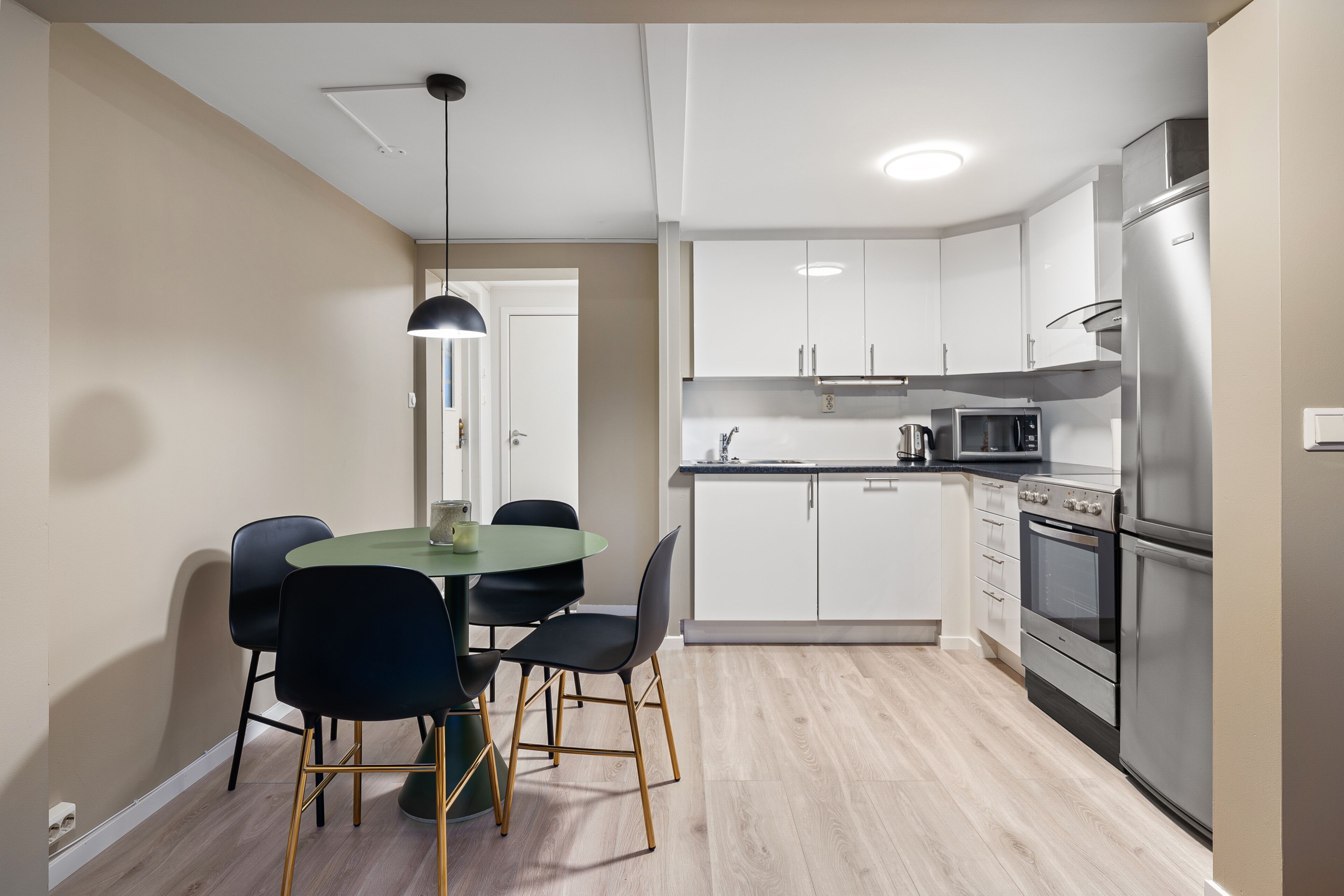 Een moderne keuken met witte kasten, een roestvrijstalen koelkast, een magnetron en een oven. Er staat een eettafel met zwarte stoelen.