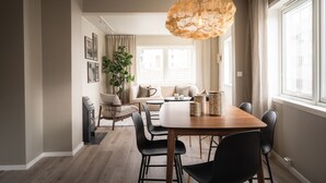Un coin repas avec une table en bois, des chaises noires et un luminaire suspendu original. On aperçoit un canapé et une plante au fond.