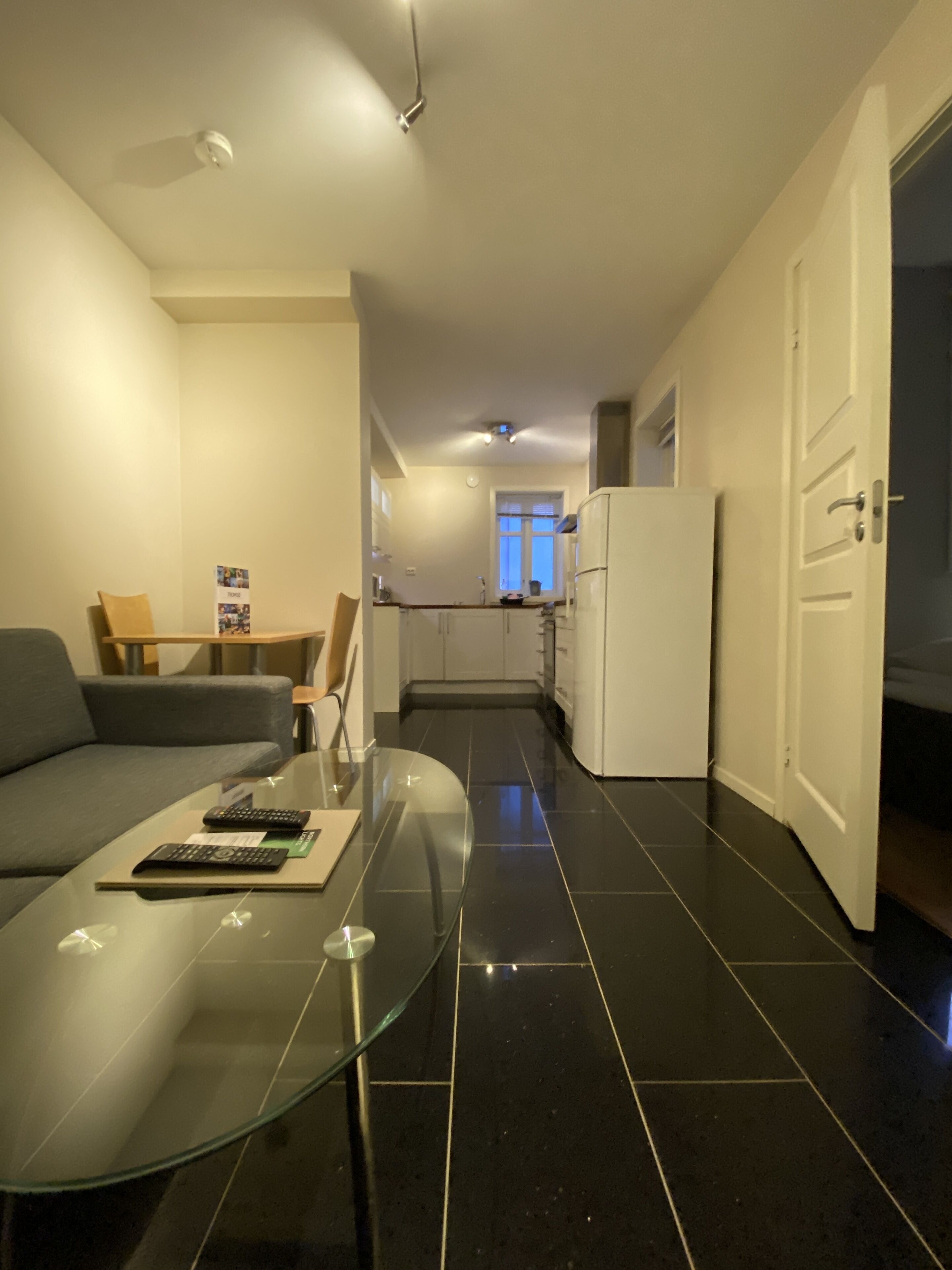 Een moderne woonkamer met een glazen salontafel, een bank en een eethoek met een tafel en stoelen. In de achtergrond is een keuken met witte kastjes en een koelkast zichtbaar.