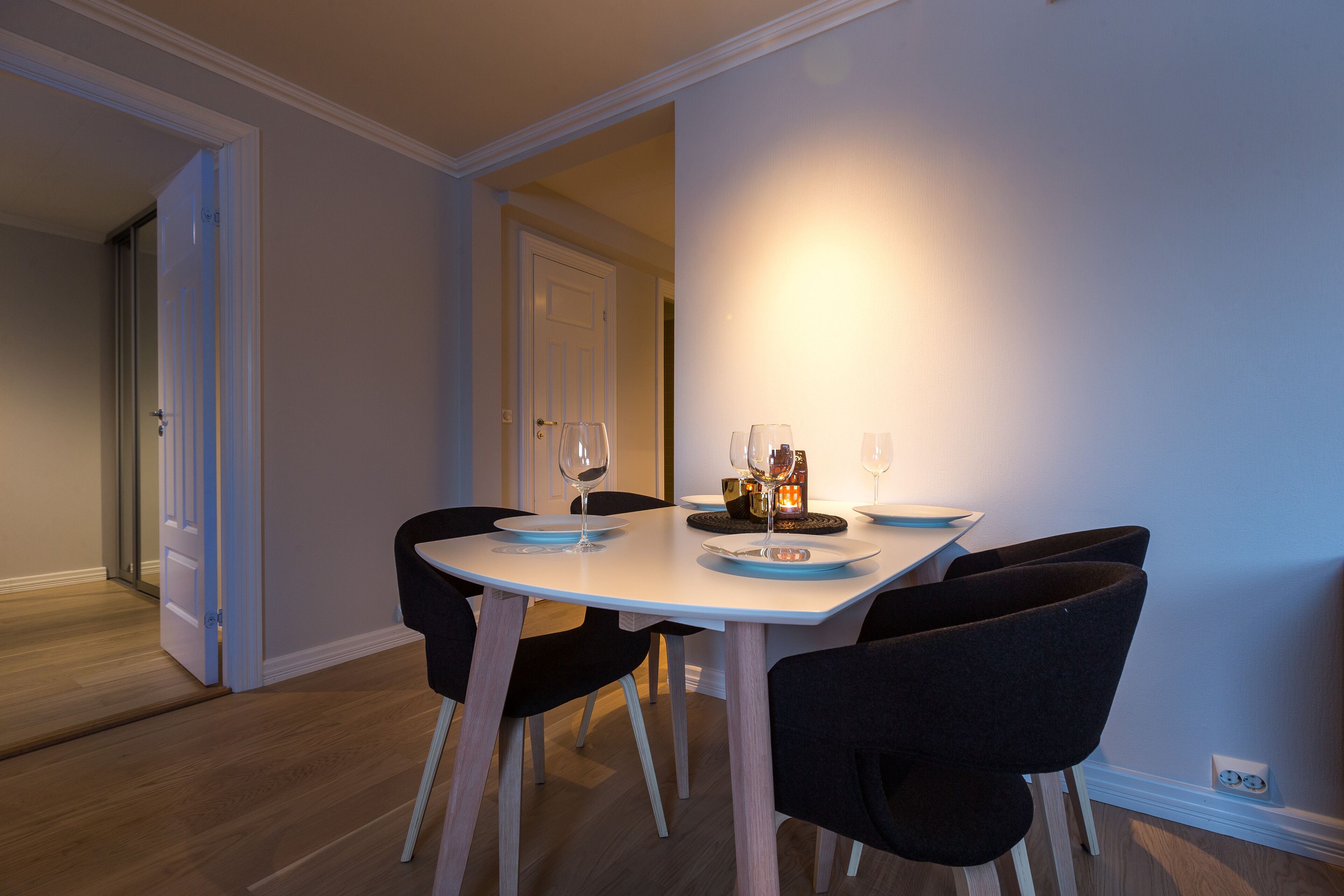 A dining area with a white table, black chairs, and a lit wall lamp.