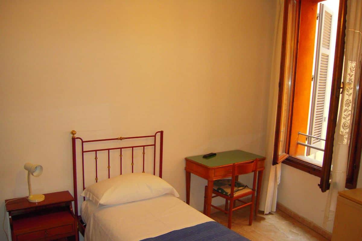 A small, simple room with a single bed, a nightstand, a small table, and a window with shutters.
