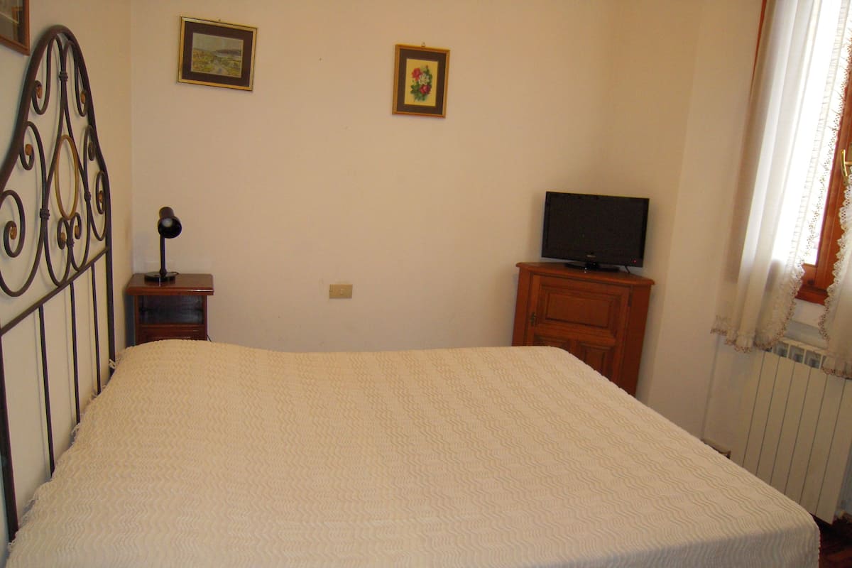 A bedroom with a bed, a television on a wooden cabinet, a small bedside table with a lamp, and two framed pictures on the wall.