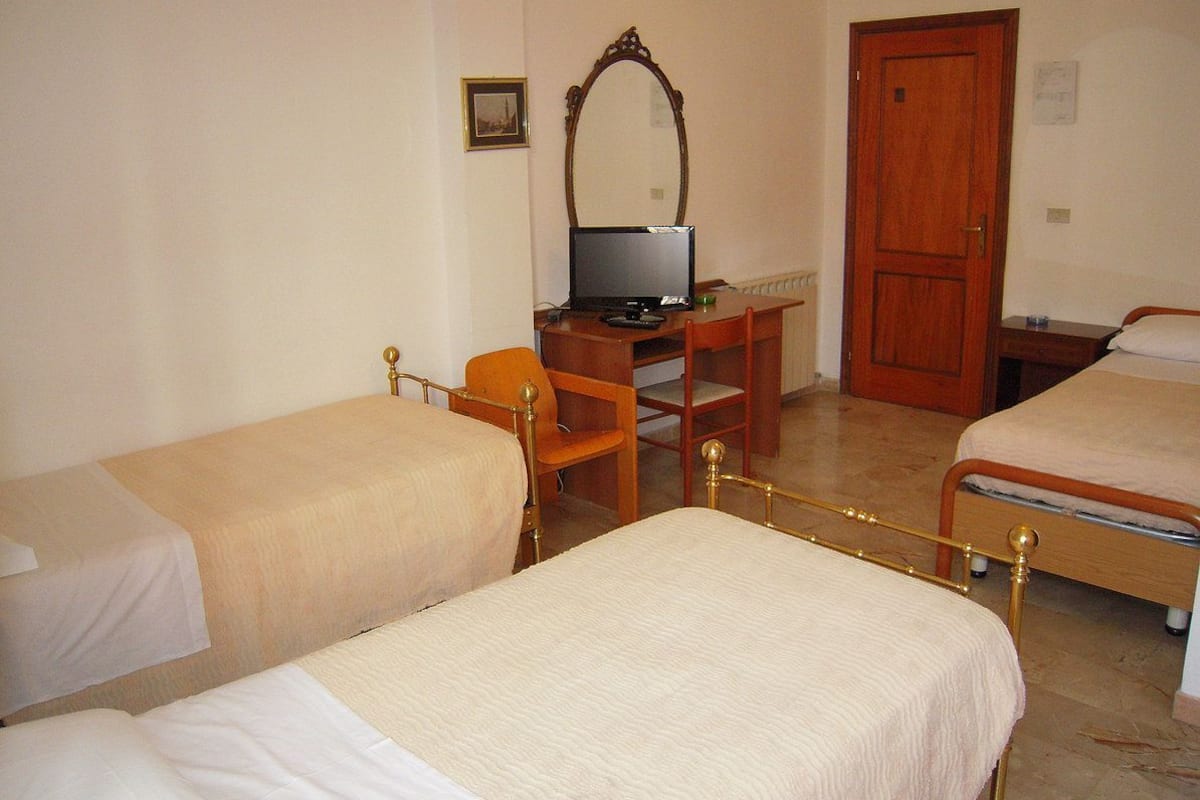 A room with two single beds, a wooden desk with a computer, a chair, and a door.