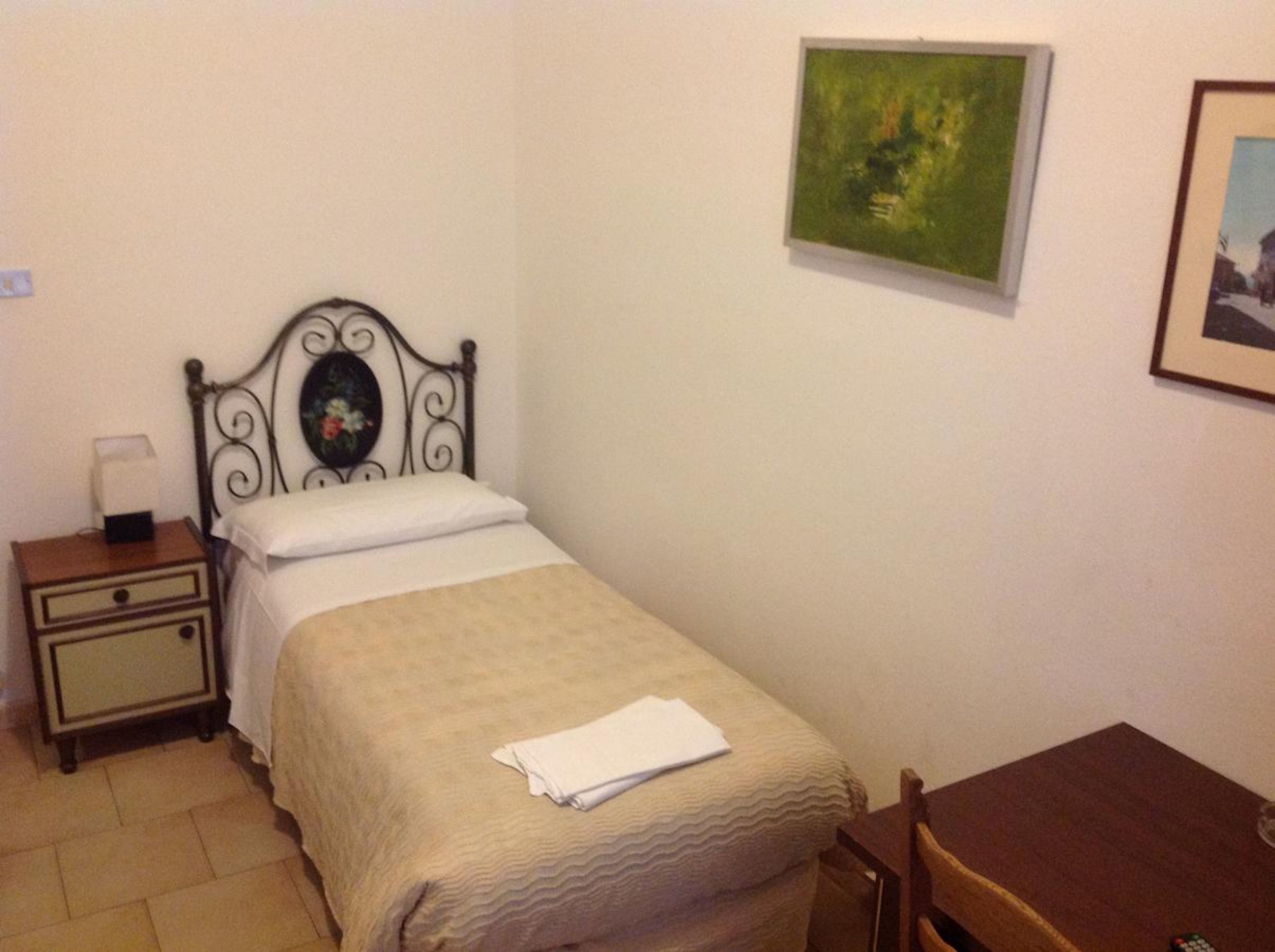 A single bed with a decorative headboard, a bedside table with a lamp, a small table with a chair, and a framed picture on the wall.