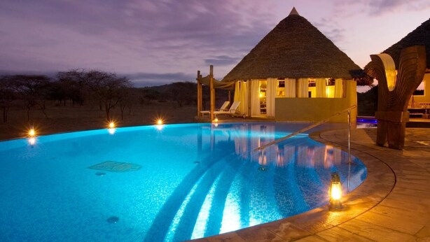 A swimming pool with a thatched-roof building adjacent to it, surrounded by a landscaped area with trees. - Severin Safari Camp (Tsavo)