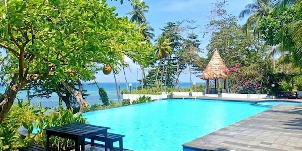 A swimming pool with a thatched-roof structure, surrounded by lush greenery and a view of the sea. - Asana Biak Papua (Biak)