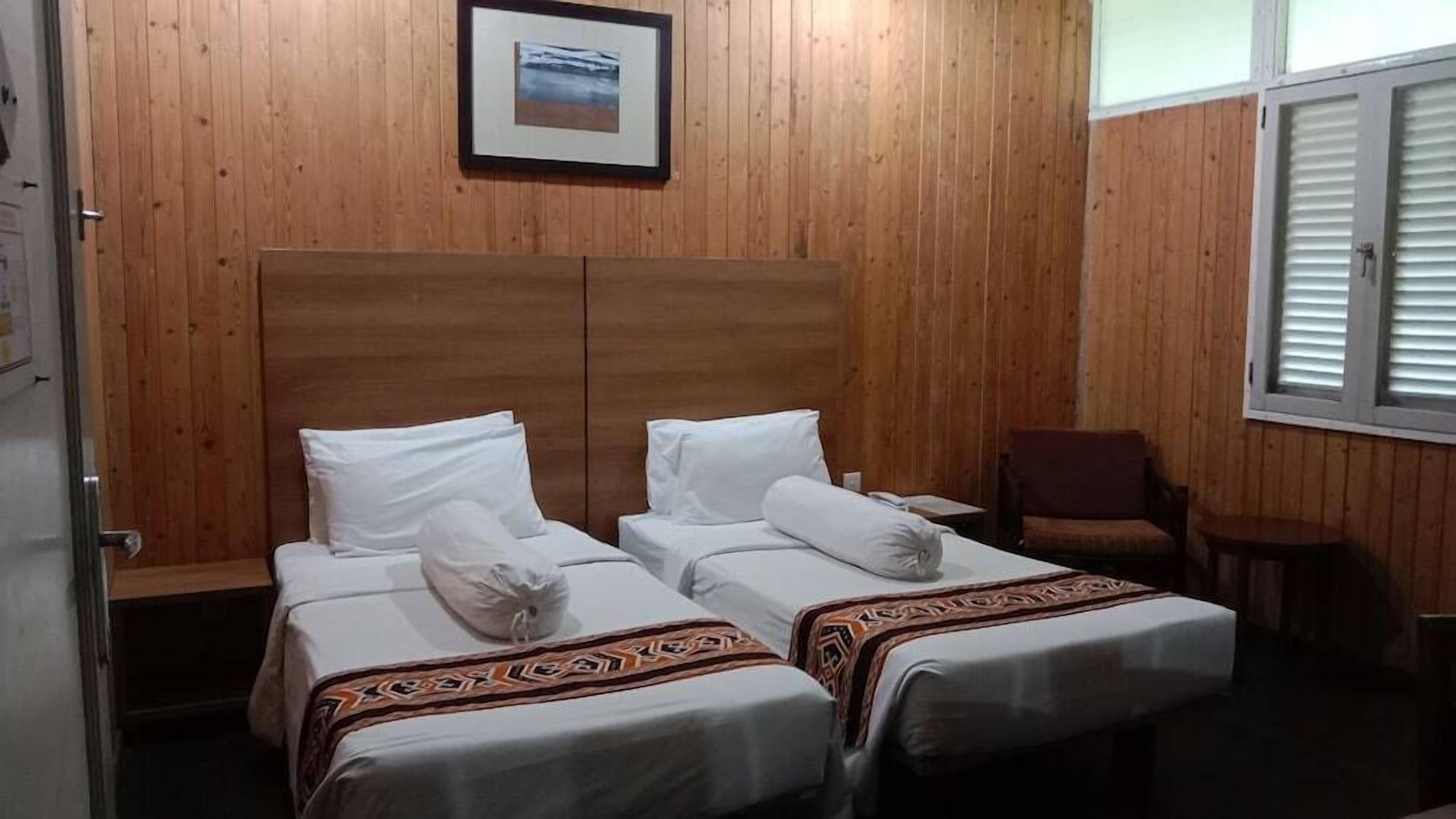 A room with two beds, wooden walls, a framed picture, and a window with blinds.