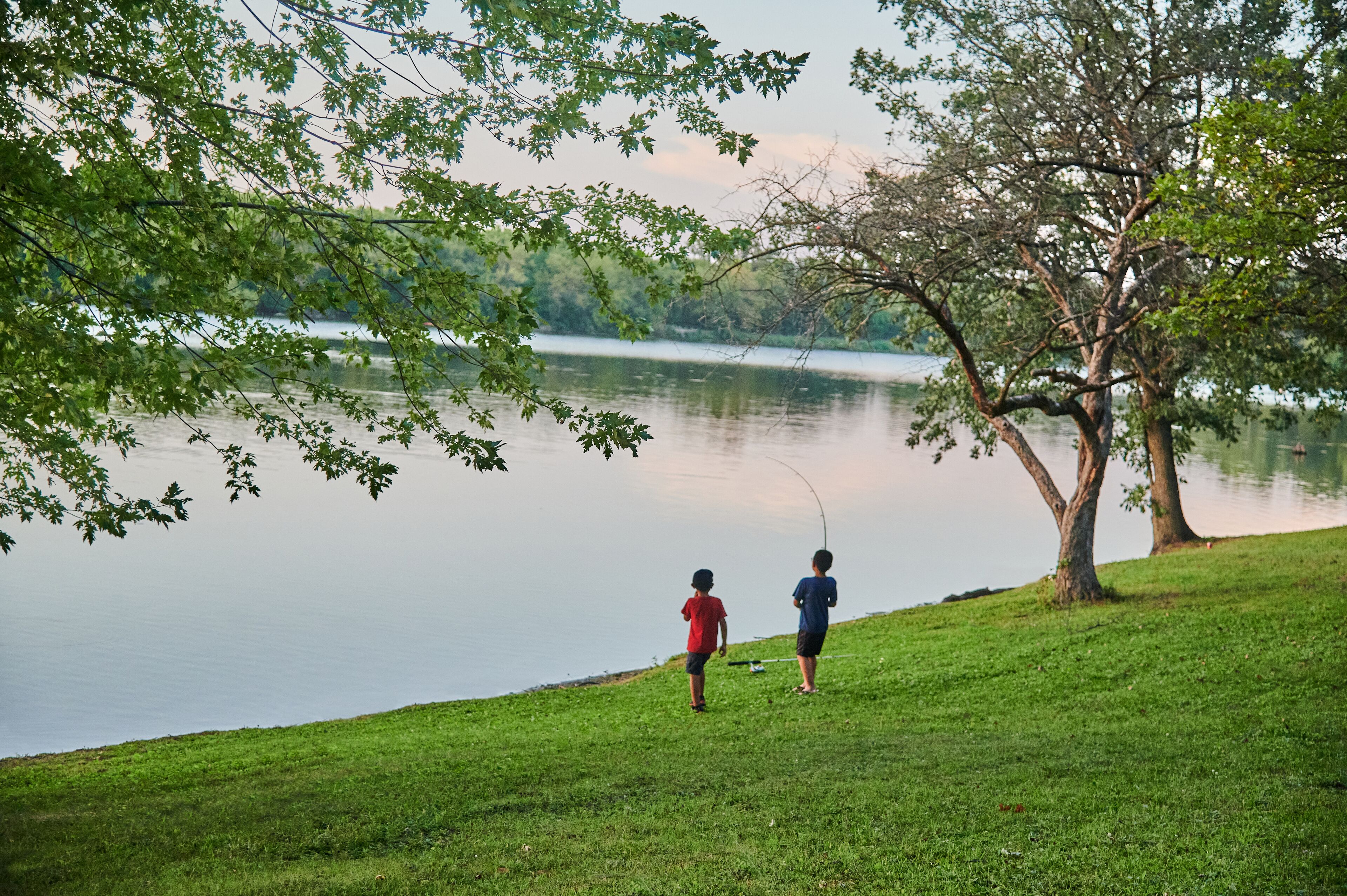 Two people fishing by a calm lake with a grassy shore and trees in the background.