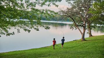 Two people fishing by a calm lake with a grassy shore and trees in the background.
