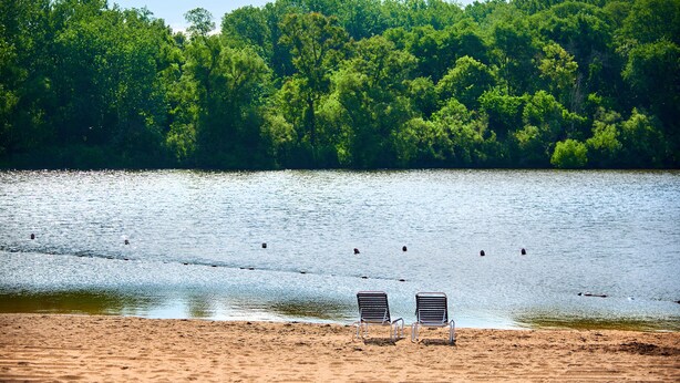 A beach with two metal chairs facing a lake surrounded by dense green trees. - O'Connell's RV Campground (Amboy)