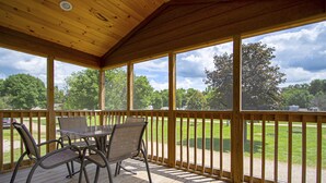 A covered outdoor area with a wooden deck, a table with chairs, and a view of a grassy field and trees. - O'Connell's RV Campground (Amboy)