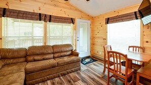 A cozy cabin interior with a wooden ceiling, a ceiling fan, a brown leather sofa, a wooden dining table with chairs, and a door with a window. - O'Connell's RV Campground (Amboy)