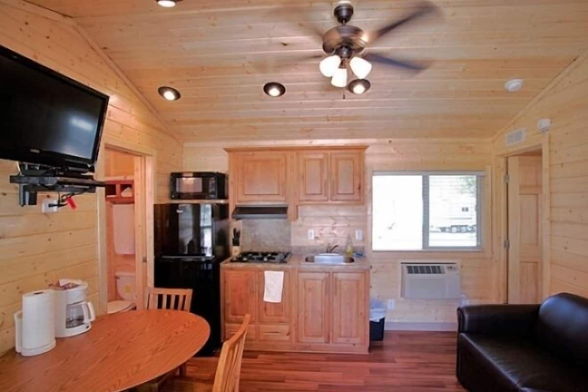 A compact cabin-style room with a wooden interior, a dining table, a sofa, a kitchen with a stove and refrigerator, and a ceiling fan.
